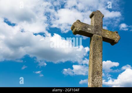 Vecchia croce gotica in pietra cristiana su sfondo blu drammatico cielo. Nuvole bianche. Particolare di antico crocifisso illuminato dal sole. Monumento d'epoca con coperchio di lichen. Foto Stock