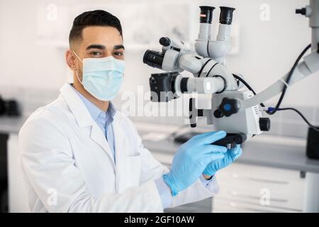 Dentista maschio arabo che indossa maschera medica protettiva usando microscopio dentale in clinica Foto Stock