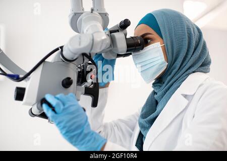 Donna dentista islamica che indossa maschera medica protettiva usando microscopio dentale in clinica Foto Stock