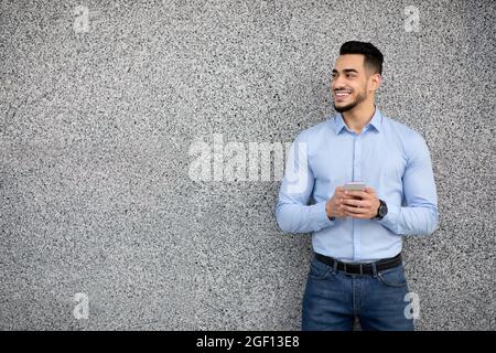 Bel ragazzo arabo che si erge su una parete grigia, utilizzando lo smartphone Foto Stock