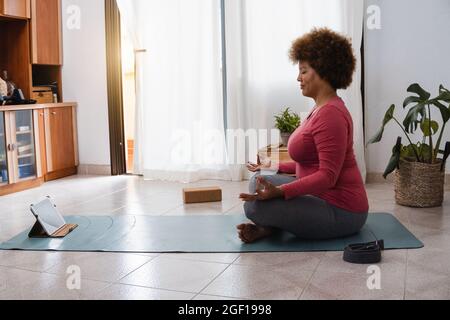 Donna africana anziana che fa yoga classe di meditazione remota a casa - Sport e stile di vita sano concetto - Focus sul tablet Foto Stock