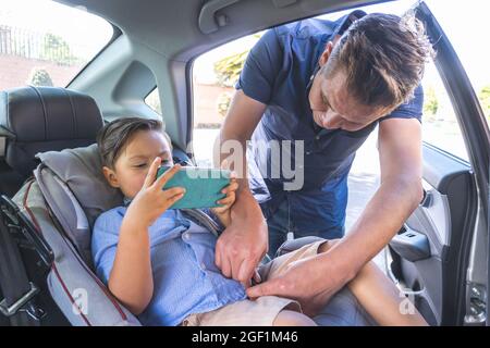 Padre che mette la cintura di sicurezza su suo figlio mentre guarda il cellulare Foto Stock