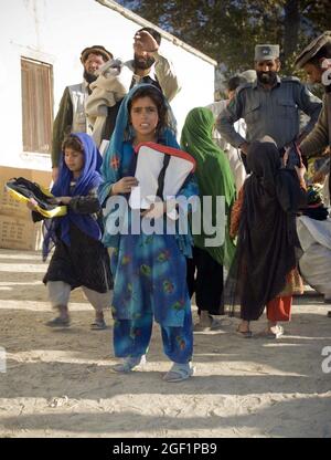 Una bambina della scuola si ferma, 4 dicembre, dopo aver ricevuto una borsa della scuola dal sub-governatore di Narang Haii Tahulam mentre un ufficiale di polizia nazionale afghano si guarda a Bar Narang, provincia di Kunar, Afghanistan. Le forniture scolastiche sono state donate da Destined Company, 2° Battaglione, 503° reggimento fanteria (Airborne). Foto Stock