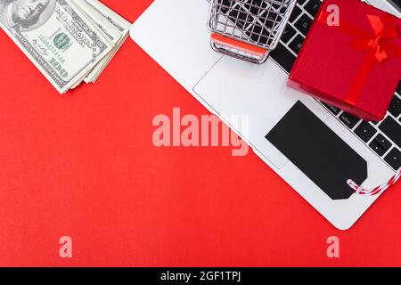Marketing online, vista dall'alto dello spazio di lavoro con tag nero vuoto, denaro, confezione regalo e shopping carrello su computer portatile su sfondo rosso e hanno copia spa Foto Stock