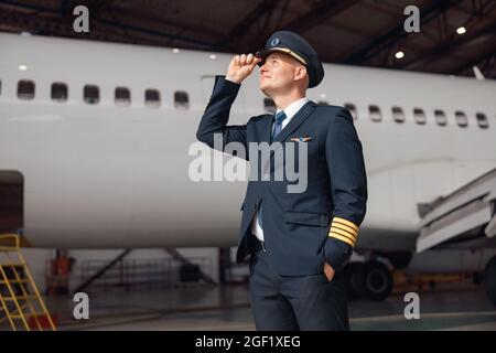 Ispirato pilota in uniforme guardando lontano, regolando il suo cappello, in piedi di fronte al grande aereo passeggeri in aeroporto hangar Foto Stock