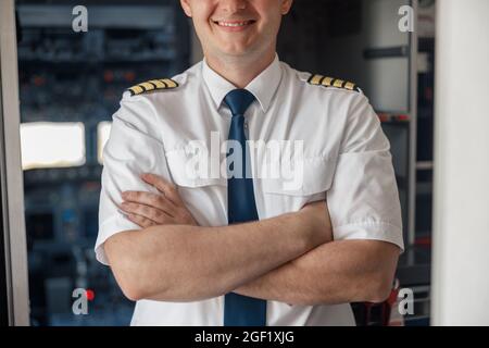 Ritratto corto di pilota maschile in uniforme mantenendo le braccia incrociate e sorridenti mentre si posa, in piedi all'interno dell'aeroplano Foto Stock