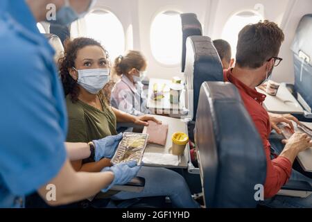 Donna con bambina che indossa una maschera protettiva che guarda l'assistente di volo femminile che serve il pranzo ai passeggeri a bordo. Viaggiare in aereo Foto Stock