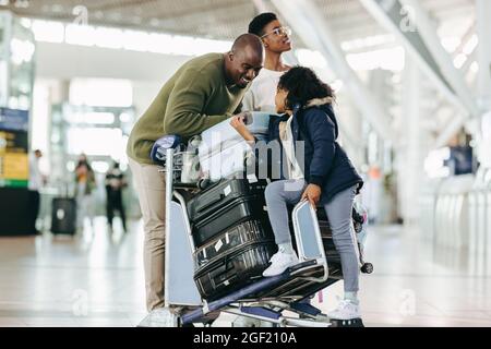 Famiglia africana con carrello bagagli in attesa al terminal dell'aeroporto per il loro volo. Padre che parla con sua figlia seduta sul trolley mentre madre che Foto Stock