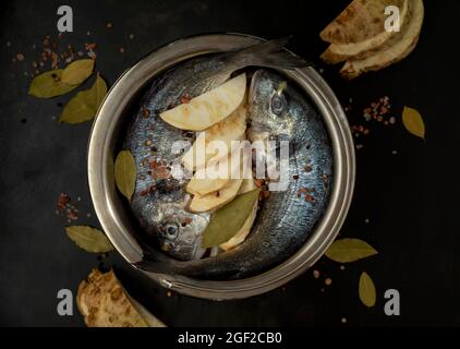 Due orate di mare crude in una pentola da cucina su sfondo nero con foglia di alloro, sale, sedano al pomolo, e pepe nero Mood Fotografia scuro Foto Stock
