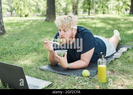 Uomo in sovrappeso sdraiato sul materassino per esercizi all'aperto e mangiare cibo sano mentre guardi qualcosa online sul laptop Foto Stock