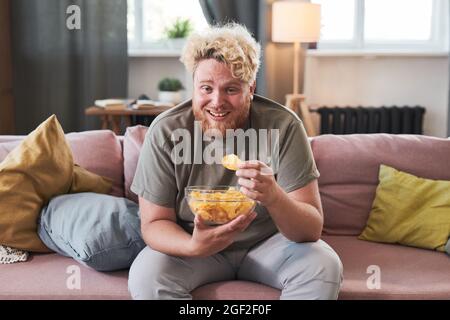 Uomo sovrappeso seduto sul divano con ciotola di patatine che li mangiano e ride mentre guarda la TV a casa Foto Stock