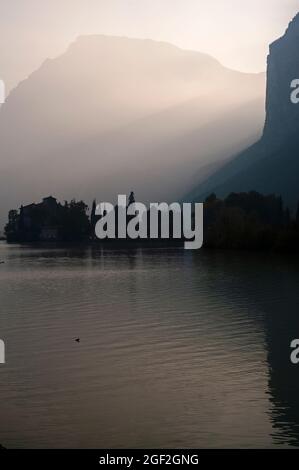 Tramonto a settembre su un lago alpino del nord Italia, il Lago di Toblino in Trentino-Alto Adige. Un castello medievale, il Castel Toblino, è in silhouette come travi dal sole affondante paesaggio aspro tra le vette dolomitiche del Monte Casale e del Monte Garzole o piccolo Dain. Foto Stock