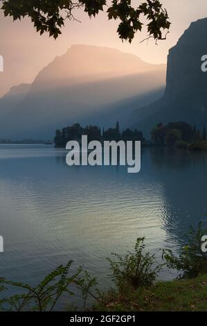 Rosa Settembre cielo serale su uno dei laghi più belli del nord Italia, il Lago di Toblino in Trentino-Alto Adige. Castel Toblino medievale, ex residenza estiva dei principi-vescovi di Trento, sorge sotto i raggi di sole che illuminano l'aspro paesaggio tra le vette dolomitiche del Monte Casale e del Monte Garzole o del piccolo Dain. Foto Stock