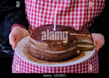 Le mani delle donne con la torta al cioccolato e una candela. Il compleanno aveva un anno. Brownie cosparse il cacao nelle mani della padrona di casa. Foto Stock