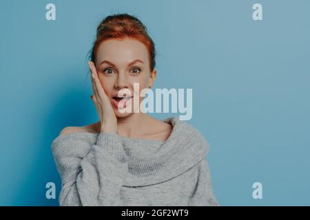 Sorpresa stupita donna rossa capelli con impressionato volto espressione isolato su sfondo blu studio Foto Stock