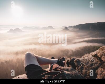 Uomo viaggiatore e turista hanno un riposo sulla cima alta montagna. Gambe maschi incrociate. Vista soleggiata di mattina presto nel paesaggio sognante. Foto Stock