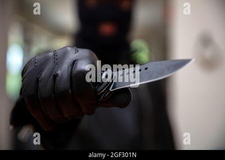 Burglar con balaclava, tenendo un coltello in mano guantata, concetto di rapina armato. Ladro minacciare con un pugnale, sfocatura sfondo interno, primo piano vista. Foto Stock