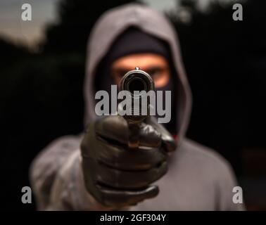 Killer accoccolato con balaclava, tenendo una pistola in mano guantata, concetto di rapina armato. Uomo che punta con una pistola alla macchina fotografica, sfondo scuro all'aperto Foto Stock