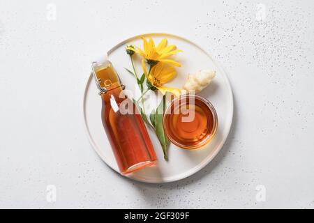 Sciroppo di carciofo di Gerusalemme in bottiglia, fiori e radice su sfondo bianco. Vista dall'alto. Dolcificanti naturali. Sostituto dello zucchero. Foto Stock