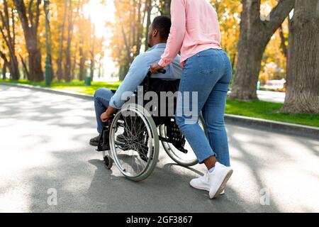 Giovane donna nera che prende il suo ragazzo paraplegico in sedia a rotelle per camminare al parco urbano in autunno Foto Stock