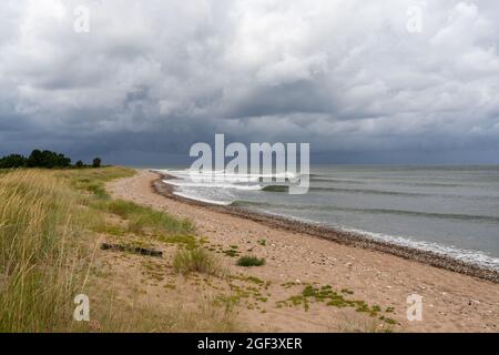 Serie di onde pulite che si infrangono su una spiaggia di sabbia con erba palude e dune di sabbia e un cielo oscuro e ominoso Foto Stock