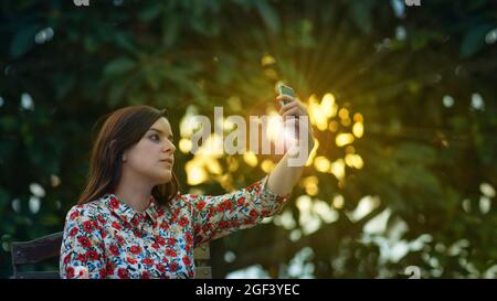 Bella giovane donna latina in abito floreale design prendendo un selfie con il suo smartphone nel giardino al tramonto con i raggi del sole che passano attraverso il Foto Stock