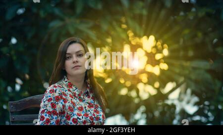 Bella giovane donna latina in abito floreale di design seduto in giardino guardando direttamente nella macchina fotografica con raggi del sole al tramonto che passa attraverso Foto Stock