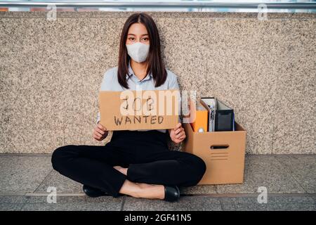 Ritratto di triste giovane donna asiatica con scatola di oggetti seduti da solo dopo essere stati licenziati dal lavoro a causa di covid-19 pandemic tenendo un cartello con lavoro voluto testo Foto Stock