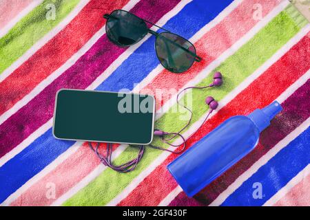 Accessori da spiaggia per vacanze estive. Vista dall'alto del telefono cellulare con schermo nero per spazio copia, auricolari, occhiali da sole, olio abbronzante, spiaggia Foto Stock