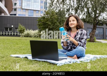 Una ragazza sorridente che siede sull'erba e usa un computer portatile. Una studentessa guarda una classe Internet e studia da remoto. La ragazza usa il wireless in Foto Stock