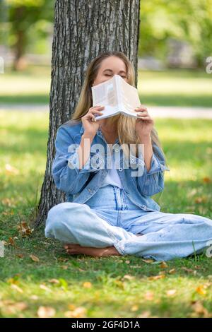 Foto verticale di una donna con un libro nel parco. Lei si siede sull'erba e ride, coprendo il viso con un libro. Concetto di riposo da gadget e c Foto Stock