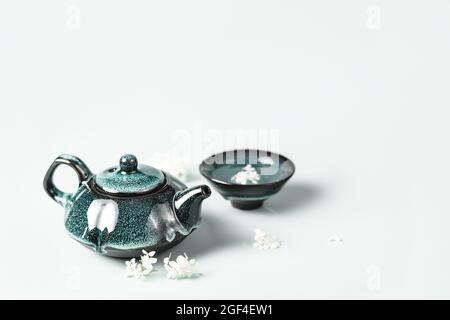 Tè verde in una teiera di argilla turchese scura e una piccola tazza decorata con fiori bianchi su sfondo pastello. Concetto creativo minimalista cerimonia del tè. Spazio di copia. Foto Stock