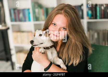 Felice donna proprietario di jack russell terrier cane, si sente la responsabilità di prendersi cura di animali domestici, in piedi contro scaffali sfondo. Persone e. Foto Stock