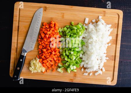 Vista dall'alto delle verdure tritate su un tagliere di bambù: Carote tritate finemente, sedano, cipolle e aglio su un tagliere di legno con coltello Foto Stock