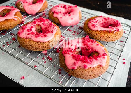 Ciambelle al forno con lampone glassa su un rack di raffreddamento a filo: Ciambelle al forno con glassa di lamponi rosa e spolverini rossi Foto Stock