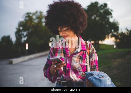 Giovane donna con capelli afro che usa smartphone all'aperto al tramonto Foto Stock