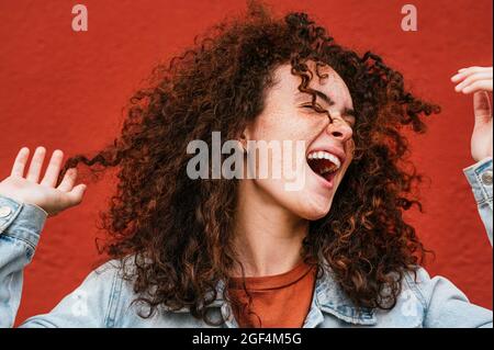 Ragazza allegra con capelli ricci che cantano davanti al muro rosso Foto Stock