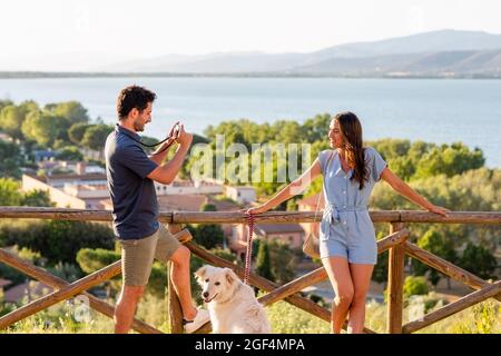 Uomo che fotografa la donna con la macchina fotografica mentre si trova in piedi con il cane da ringhiera Foto Stock