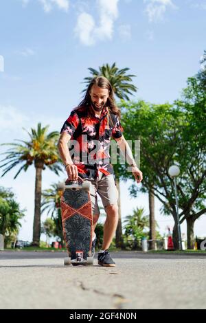 Uomo in camicia hawaiana che tiene skateboard su strada Foto Stock
