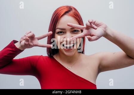 Sorridente donna rossa che fa segno di pace di fronte a sfondo bianco Foto Stock