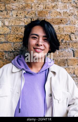 Giovane uomo sorridente che indossa una giacca con cappuccio davanti al muro di mattoni Foto Stock
