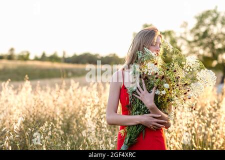 Giovane bionda donna che odora fiori selvatici bouquet in campo Foto Stock