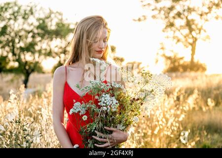 Bella donna in abito rosso che tiene fiori in campo Foto Stock