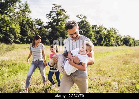 Sorridendo padre che porta figlio mentre cammina con la famiglia sul prato Foto Stock