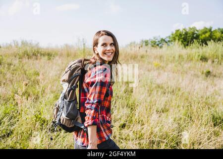 Donna escursionista sorridente mentre si guarda oltre la spalla in giorno di sole Foto Stock