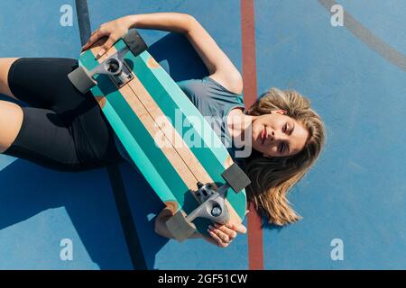 Donna bionda con skateboard sdraiata al campo di pallacanestro durante la giornata di sole Foto Stock