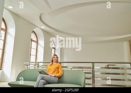 Donna d'affari bionda seduta con tablet digitale sul divano nella lobby dell'ufficio Foto Stock