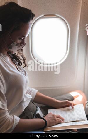 Donna d'affari che legge libro mentre viaggia in aereo durante COVID-19 Foto Stock