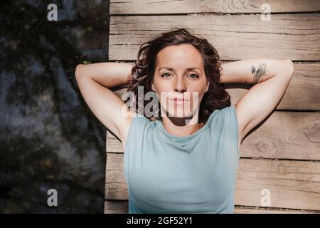 Donna adulto medio con le mani dietro la testa che giace sul molo vicino al lago nella foresta Foto Stock