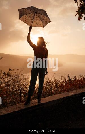 Silhouette di donna con ombrello in piedi sul muro di ritegno al tramonto Foto Stock